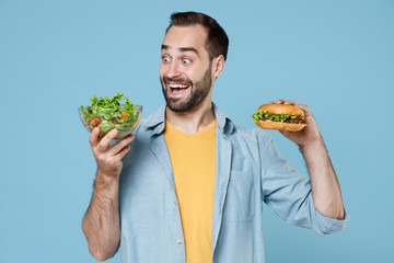 Excited surprised young bearded man guy 20s wearing casual clothes posing hold vegetable salad in glass bowl american fast food burger isolated on pastel blue color wall background studio portrait.