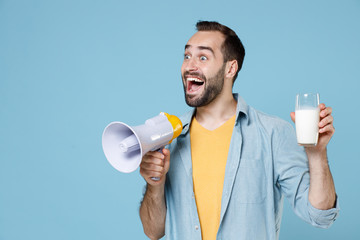 Surprised young bearded man guy 20s wearing casual clothes posing screaming in megaphone holding in hand glass of milk looking aside isolated on pastel blue color wall background studio portrait.