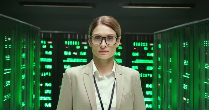 Portrait shot of young serious Caucasian pretty woman in glasses, worker of database storage looking at camera. Female engineer or analytic in datacenter monitoring room. dolly shot.