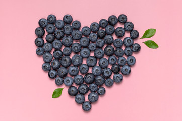 Fresh juicy blueberries with green leaves in the shape of heart on pink background. Blueberries background. Flat lay top view. Creative love concept. Healthy berry, organic food, antioxidant, vitamin