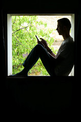 Person using technology. Silhouette of a man in the inset of a window, with a smartphone in his hands. Background deliberately blurred with vegetation in the sun.