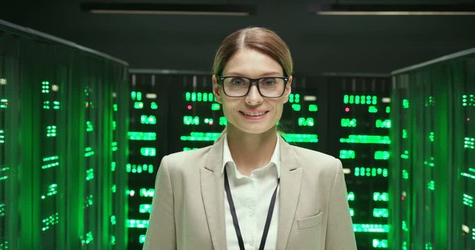 Portrait shot of young serious Caucasian pretty woman in glasses, worker of database storage turning face and smiling at camera. Close up of female engineer or analytic in datacenter monitoring room.