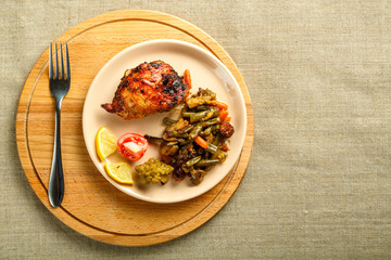 A plate with vegetables and a piece of chicken baked on a round stand on a wooden table with a fork.