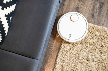 White robotic vacuum cleaner cleans the living room on the floor against the backdrop of gray sofa...