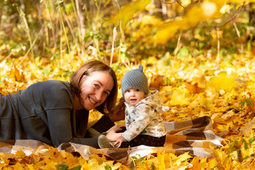 Young mother spending time with her baby in autumn park in sunny day. They are sitting on yellow leaves happily smiling