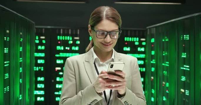 Caucasian young beautiful woman in glasses standing at servers and tapping or scrolling on mobile phone. Female analytic in data storage texting message on smartphone. Digital security of database.