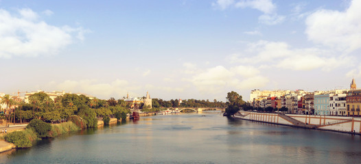 Obraz premium Landscape panorama of Seville with the Guadalquivir river, Triana, the bridge and the city surrounded by palm trees, and a blue sky in the background for a postcard or a wallpaper