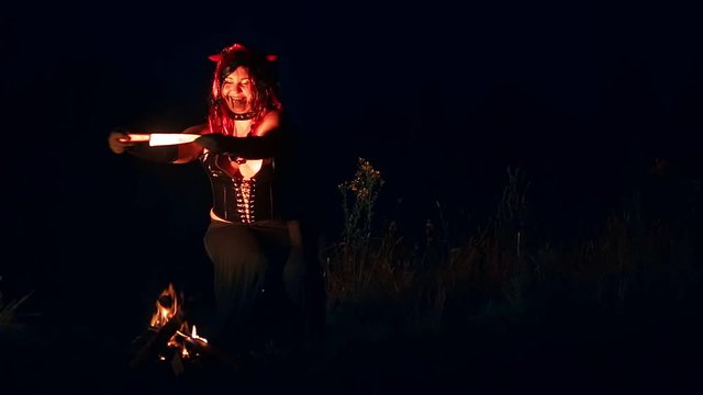 A woman in the form of a devil in black with horns at night by the fire with a knife before the sacrifice