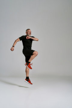 Full length shot of cheerful middle aged muscular man in black sportswear jumping while having workout in studio over grey background