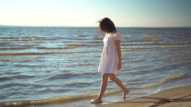 Attractive young woman walks along the sea beach. A girl in a white dress walks barefoot along the beach.