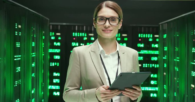 Portrait of beautiful Caucasian young woman tapping and scrolling on tablet device in big data storage room and smiling to camera. Female system administrator in glasses using gadget computer.