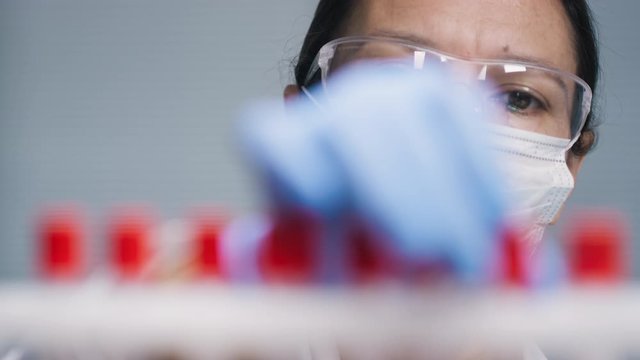 Tilt up close up shot of female biomedical scientist in goggles and face mask putting test tubes with blood samples from patients tested positive for covid-19 in rack