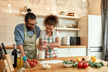 Young couple in love preparing the dough for making pizza with vegetables at home. Man and woman wearing apron, cooking together indoors. Hobby, lifestyle