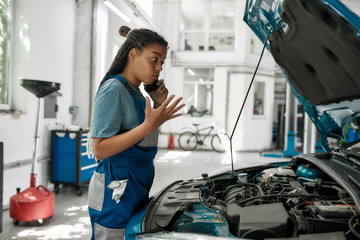 Personalized service. Young african american woman, professional female mechanic talking on phone emotionally, while examining engine under hood of car at auto repair shop