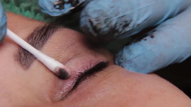 beautician is using small cotton swab for remove dye from eyelids of female client in a beauty salon.