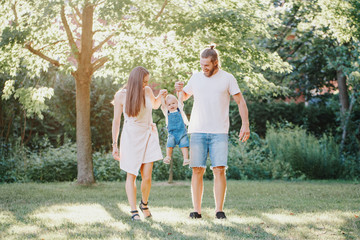 Naklejka na ściany i meble Happy smiling mother and father with baby boy in park outdoors. Family Caucasian mom and dad with son walking hugging in park on summer day. Happy family authentic lifestyle concept.