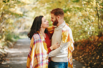 Beautiful couple man woman in love. Smiling boyfriend and girlfriend wrapped in yellow blanket hugging in park on autumn fall day. Togetherness and happiness. Authentic real people outdoors.