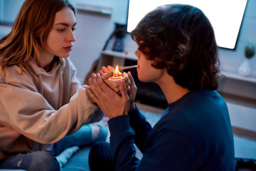 Young girl lighting cannabis cigarette for her boyfriend while sitting on the couch at home. Young...