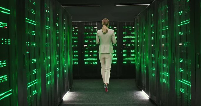 Rear on Caucasian beautiful woman walking away among servers with tablet device, checking big data processors. Back view on female analytic in data storage working on gadget over digital security.