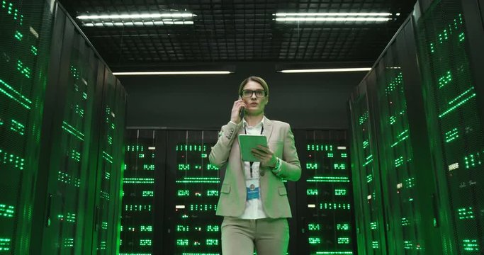 Caucasian young woman in glasses walking at servers with tablet gadget in hands and talking on mobile phone. Female engineer in data storage speaking on cellphone about digital security of database.