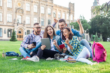 Group of excited young people celebrating success while sitting outdoors on grass lawn in the park with tablet computer