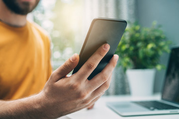 Cropped shot of smiling male blogger sitting at home at the working desk browsing internet on his mobile phone checking his new post comments from his foolowers in social network application