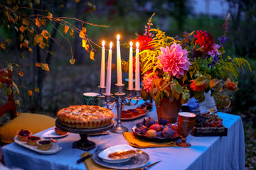 Autumn evening photo shoot - romantic dinner outdoors. Table with tablecloth and decoration - pie, figs, glasses, plates, table setting and candelabra with candles. Fall flowers dahlia bouquet