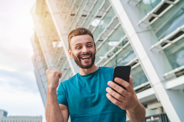 Guy celebrating a bet win in online sport gambling application on his mobile phone clenching his fist in winner’s gesture