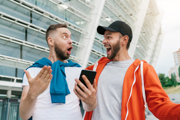 Two friends looking extremely excited getting good news about winning a bet in online bookmaker watching broadcast with winner results on mobile phone with football stadium on the background