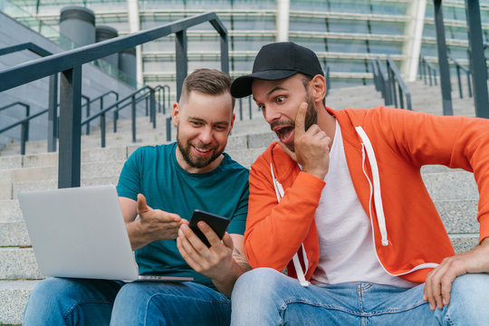 Two lucky friends sitting on football stadium steps and making bets using online sport betting services on their mobile phone and laptop computer