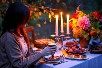 Autumn evening photo shoot: young woman with wine glass at romantic dinner in twilight nature outdoors. Table decoration - cake, figs, plates, candelabra with candles, fall flowers. Red dahlia bouquet