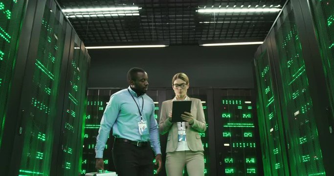 Caucasian woman walking passage with servers in datacenter and talking with African American man engineer while showing information on tablet device. Multiethnic digital security cryptographers.