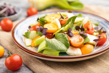 Vegetarian salad of pac choi cabbage, kiwi, tomatoes, kumquat, microgreen sprouts on gray wooden background. Side view, selective focus.