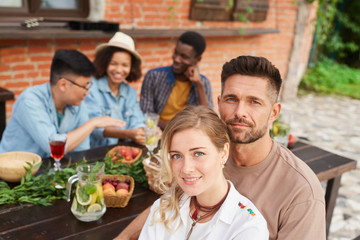 Group of beautiful young people enjoying dinner outdoors in sunlight, focus on smiling couple in foreground, copy space