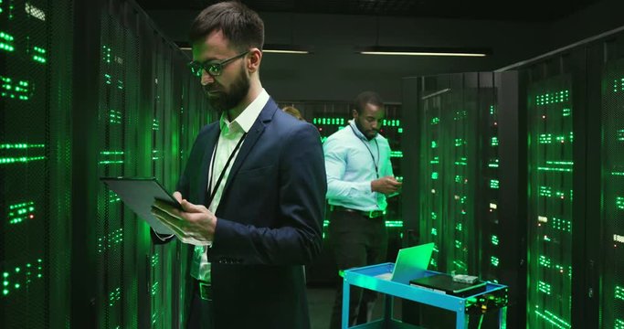 Caucasian man technician in database room with servers, tapping on tablet device and coding information. Male security worker in datacenter using gadget. African American cryptograph on background.