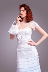 Portrait of Sad Redhaired Caucasian Female Posing in White Wedding Dress and Little Purse Against Gray Background.