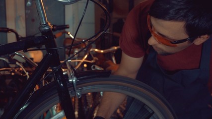 Fototapeta na wymiar theme small business bike repair. Young brunette Caucasian man in protective glasses, gloves and fartuhe uses hand tools to repair and tuning Rim Brakes and wheel spinning bike in a garage workshop