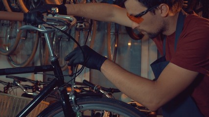 Obraz na płótnie Canvas theme small business bike repair. Young brunette Caucasian man in protective glasses, gloves and fartuhe uses hand tools to repair and tuning Rim Brakes and wheel spinning bike in a garage workshop