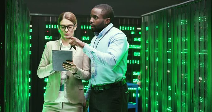 Multiethnic man and woman talking and checking database storage with servers. Using tablet device. Couple of mixed-races colleagues discussing security. Male and female analytics cryptographers.