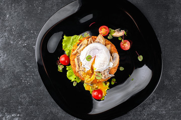 Fried beefsteak with mushrooms, poached egg and tomatoes on a black plate, black background