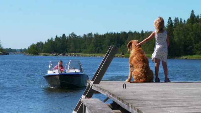 A blonde young girl and her dog wait for her dad return by boat on a pier