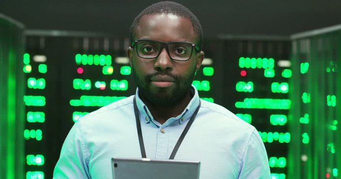 Close up of handsome African American cheerful young man in glasses holding tablet device and smiling in big data storage room of processors. Male system administrator with gadget computer at servers.