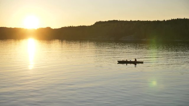 silhouette of person with little child sailing small kayak along large tranquil lake against dark forest at bright sunset