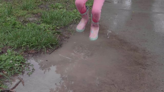 Young Caucasian girl wearing pink rain boots and striped purple shirt jumps up and down in puddle splashing water, handheld slow motion