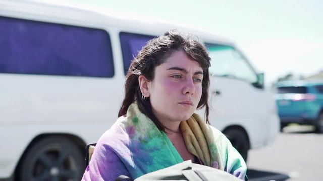 Dark-haired girl wrapped in colorful towel stands by white van, windy