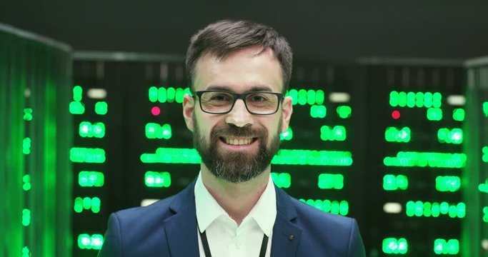Portrait of Caucasian handsome happy man in glasses turning face to camera and smiling among servers with big data information. Green working processors in database storage. Close up of cheerful male.