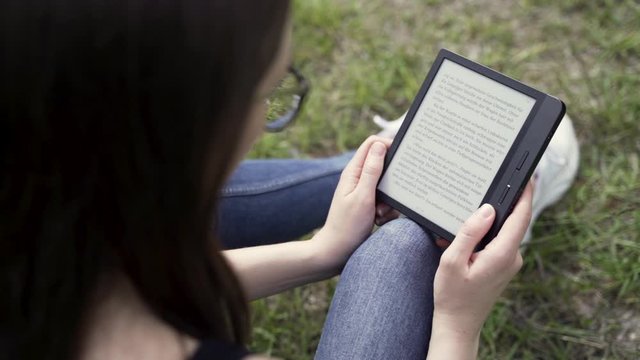 Young student girl with glasses reads book on e-reader in nature, overshoulder