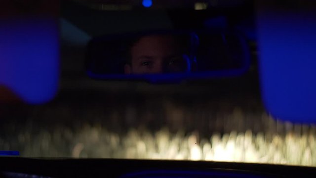 A young Caucasian male, nervously watching through his rearview mirror, during a traffic stop at night. Slowed to half speed.