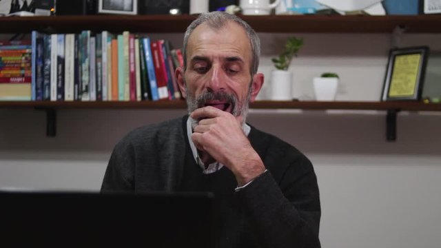 Mature man working from home on his laptop thinks and hesitates stroking his beard