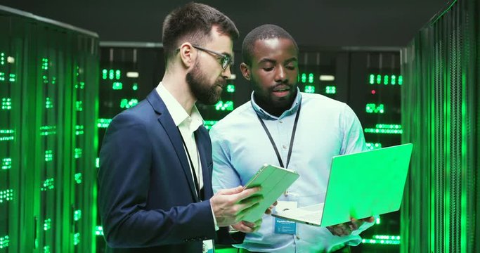 Multiethnic men talking and checking database storage with servers. Using tablet device and laptop computer. Mixed-races colleagues discussing security. Males analytics cryptographers.